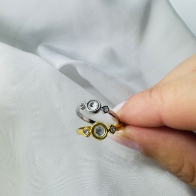 Dois anéis, um prateado e outro dourado, com pedras transparentes, segurados por uma mão sobre tecido branco