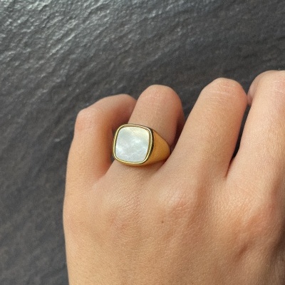 Anel dourado com pedra branca quadrada em mão sobre fundo preto texturizado