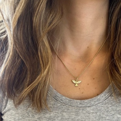 Colar dourado com pendente de anjo usado no pescoço de mulher com camiseta cinza.