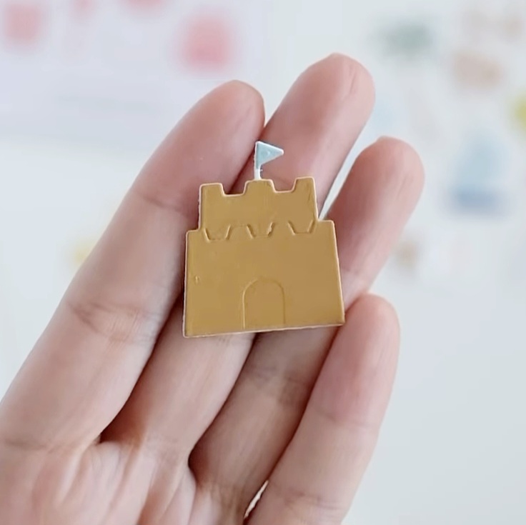 Pin castelo de areia amarelo com bandeira azul clara na mão