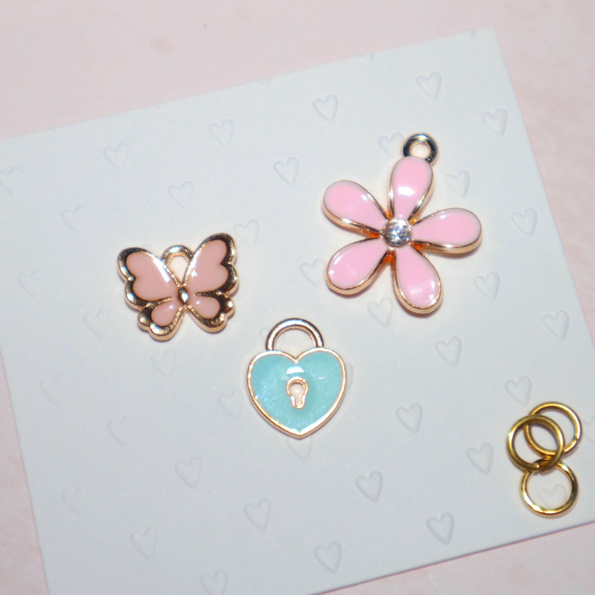 Três pendentes esmaltados coloridos e três argolas douradas, sobre papel com corações em relevo.
