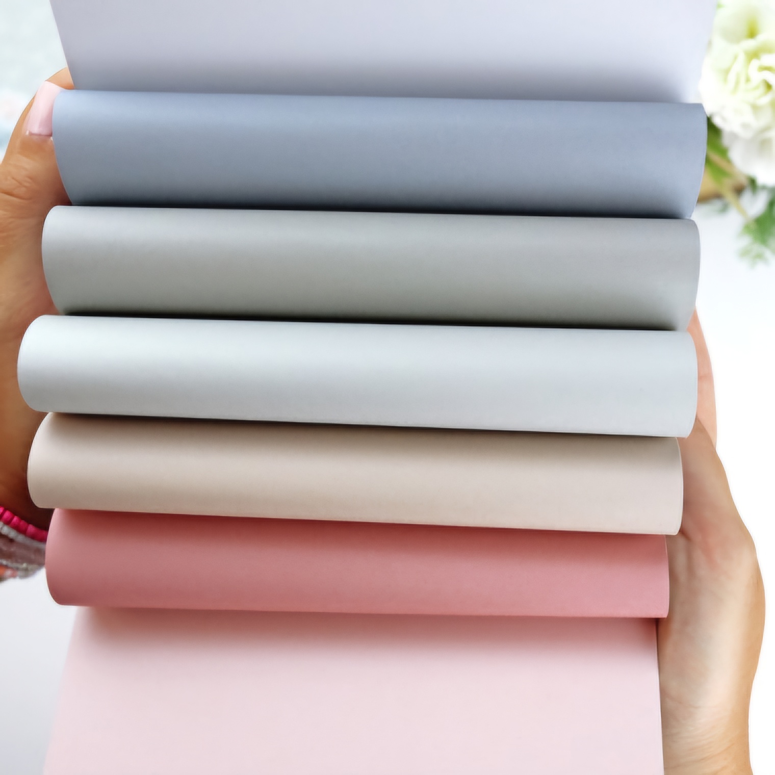 Seis rolos de tecido em várias cores neutras segurados por mãos com fundo desfocado