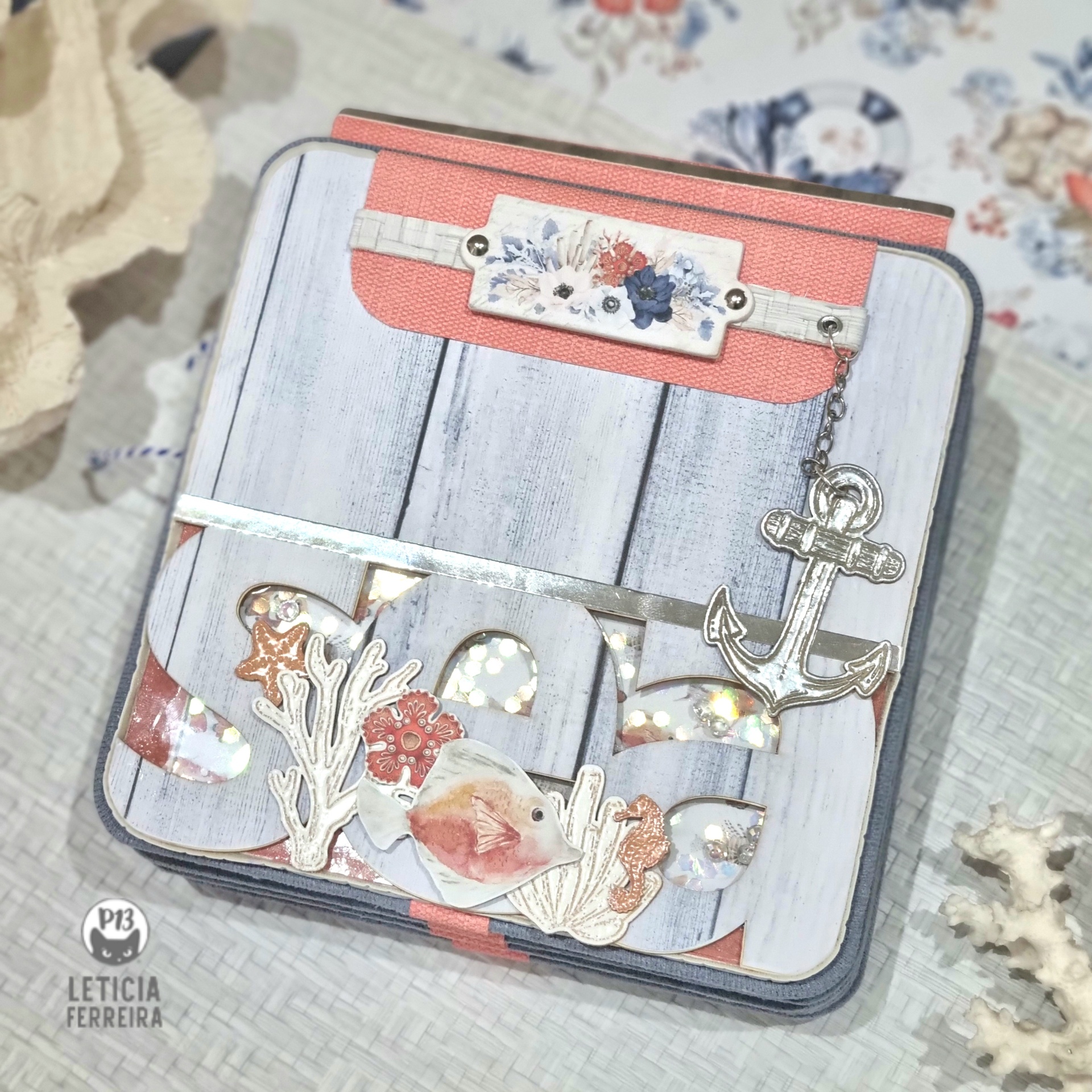 Estojo quadrado com padrão de tábuas brancas e detalhes em coral, com enfeites metálicos de anzol e corais.