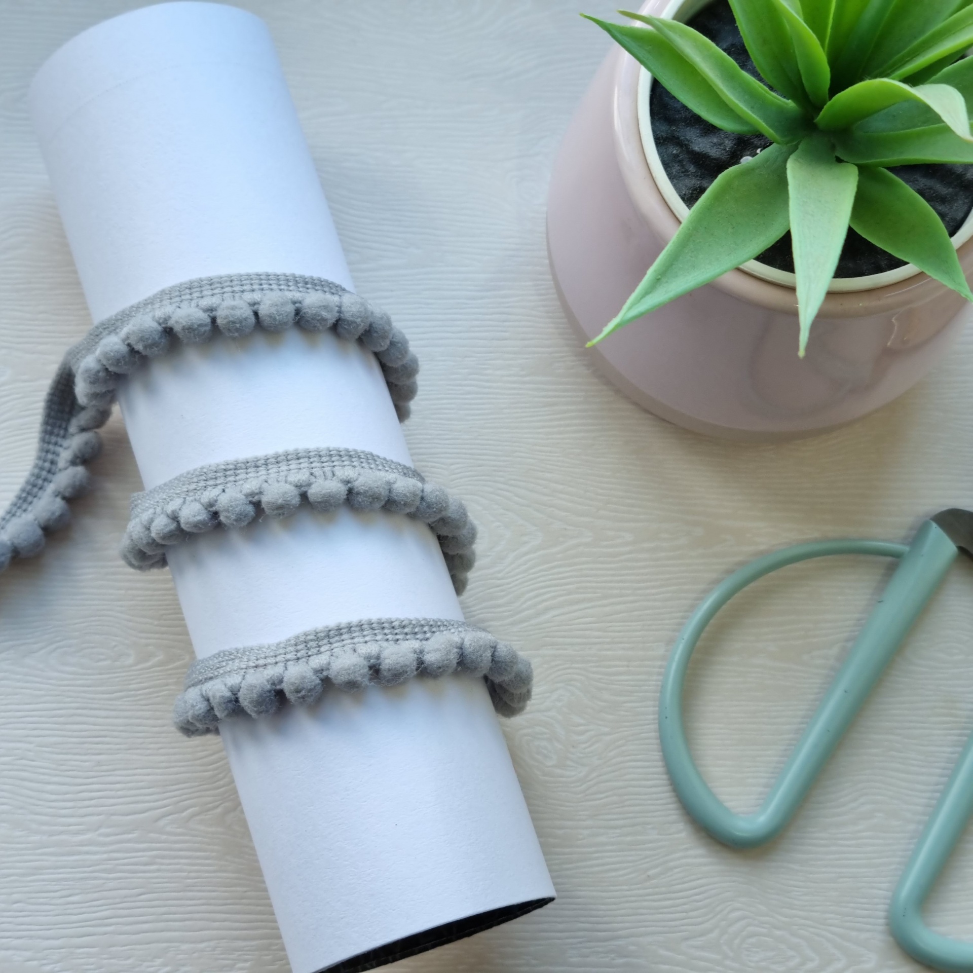 Fita decorativa cinzenta com bolinhas enrolada num rolo branco junto a vaso com planta e utensílio verde
