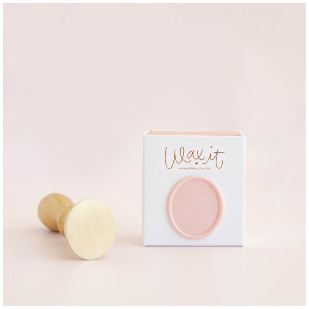 Selo de metal com cabo em madeira ao lado de uma caixa branca com etiqueta rosa e texto 'Wax it'