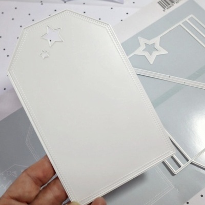 Cartão branco em forma de etiqueta com estrelas recortadas e moldes de corte metálicos ao fundo