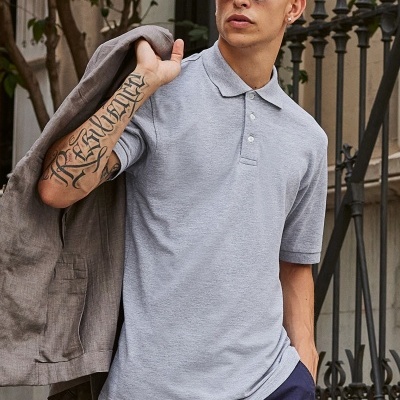 Homem jovem com polo cinzento claro, calças azul-escuras e casaco cinzento claro, usa óculos escuros e tem tatuagem no antebraço.