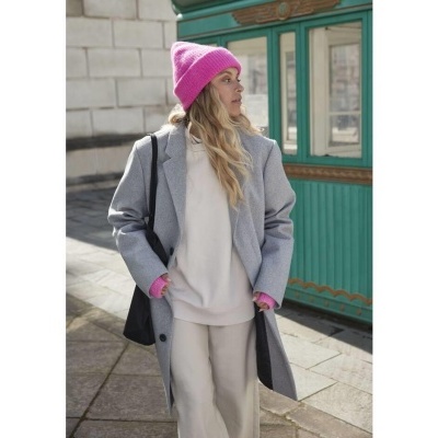 Mulher com casaco cinza, camisola e calças bege, gorro e luvas cor-de-rosa, bolsa preta, em rua com estrutura verde de fundo.