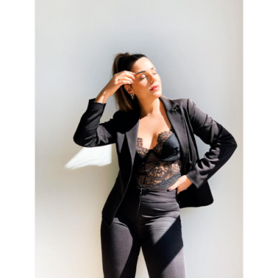 Mulher com blazer preto, calças pretas e body renda preto contra parede branca