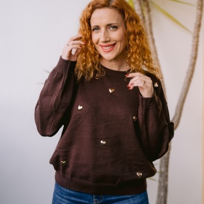 Mulher a usar camisola castanha com corações dourados e jeans azuis em interior com planta.