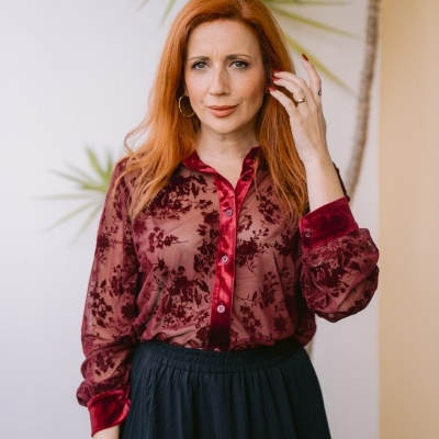 Mulher vestindo camisa bordô translúcida com padrão floral e saia preta em ambiente com plantas ao fundo