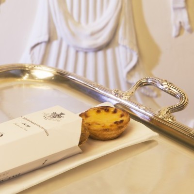 Pastel de nata em embalagem branca sobre bandeja prateada com fundo decorativo branco.