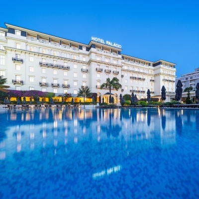 Hotel clássico branco com piscina azul e sombrinhas pretas em frente.