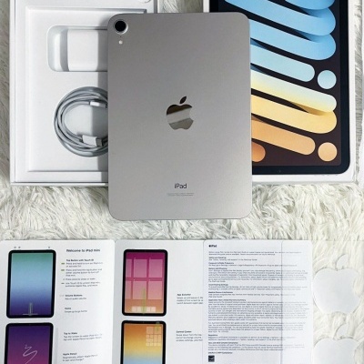 iPad cinzento prateado com caixa aberta, cabo USB e manuais num fundo branco texturizado