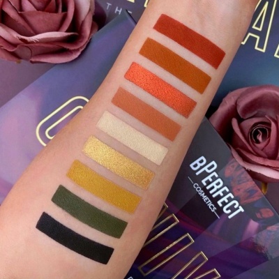 Amostras de sombras de olhos em tonalidades diversas no braço, embalagem BPERFECT COSMETICS e flores cor de rosa