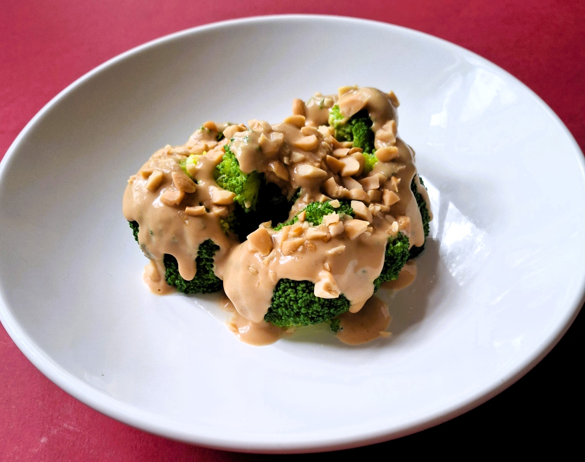 Brócolos c molho de amendoim Brócolos com molho e nozes num prato branco sobre fundo vermelho