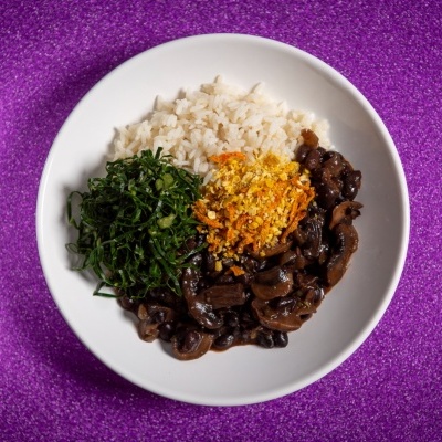 Prato branco com arroz, couve, farofa e carne com molho em fundo roxo brilhante