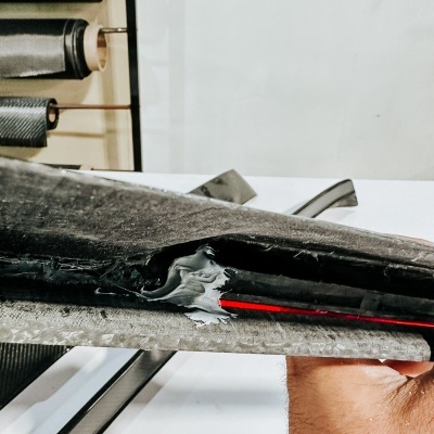 Prancha de surf preta danificada com camada vermelha visível, mão segurando-a, fundo com rolos e autocolantes