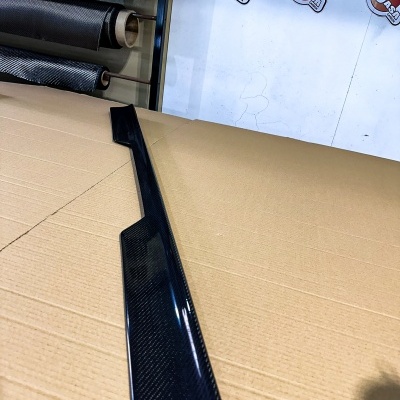 Peça automóvel em fibra de carbono preta sobre cartão com fundo de rolos e painel com texto JASA