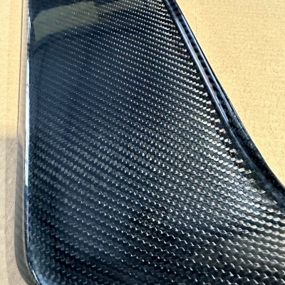 Peça preta em fibra de carbono com padrão tecido sobre superfície castanha com texto e URL