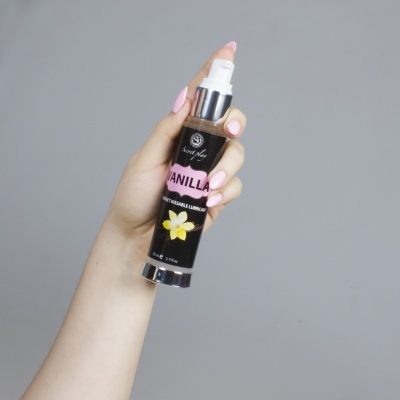 Frasco spray preto com texto VANILLA e flor amarela, mão segurando, fundo cinza