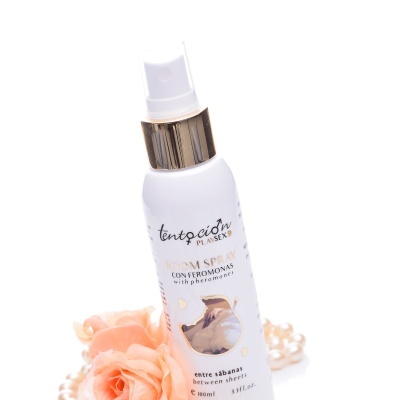 Frasco branco de spray com texto e tampa dourada, rodeado por flores rosas e pérolas.