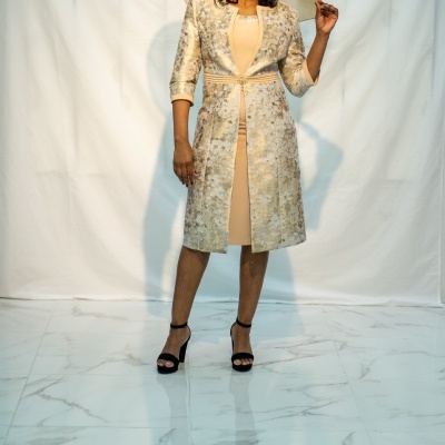 Mulher com vestido bege e casaco floral dourado, chapéu bege e sandálias pretas de salto alto