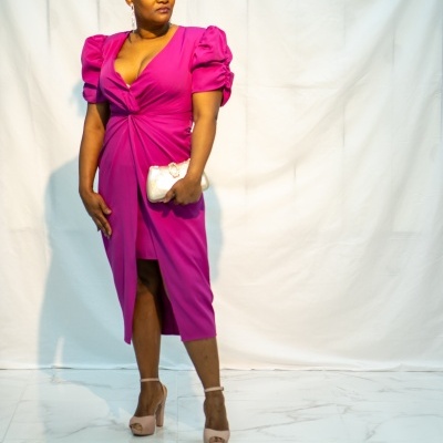 Mulher veste vestido fúcsia com mangas bufantes e segura carteira dourada em fundo branco.