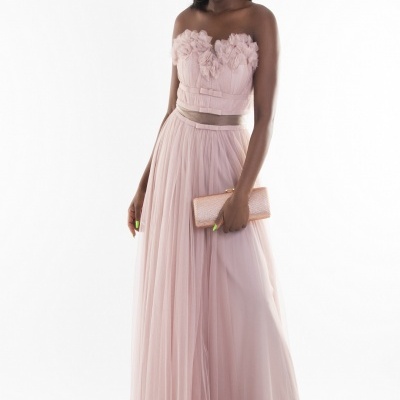 Vestido de festa rosa claro com flores no corpete e saia longa esvoaçante, modelo segurando carteira rosa em fundo branco