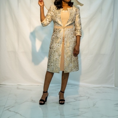 Mulher com vestido e casaco bege com padrão floral dourado, chapéu bege decorativo e sandálias pretas de salto alto