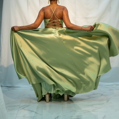 Vestido longo verde acetinado com costas cruzadas, visto de costas num piso de azulejos brancos.