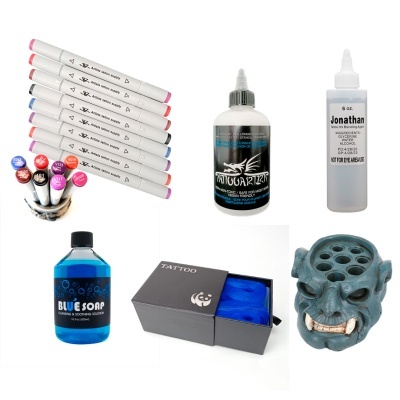 Conjunto de suprimentos para tatuagem incluindo marcadores, tintas, sabão, caixa com tecido azul e suporte de cerâmica em forma de cabeça.