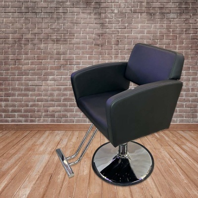 Cadeira de salão preta com base cromada, sobre chão de madeira e muro de tijolos vermelhos.