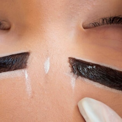 Pessoa com produto preto nas sobrancelhas e aplicador em mão com luva branca