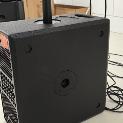 Alto-falante preto com suporte metálico no topo e cabos pretos num espaço com chão claro e parede de azulejos brancos