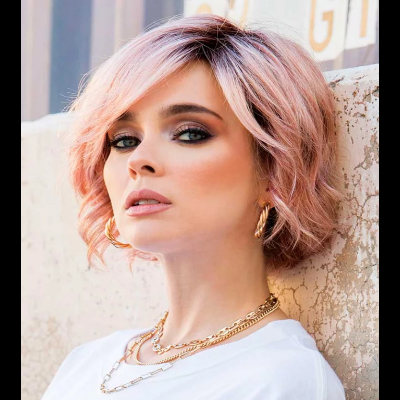 Mulher com cabelo rosa claro e joias douradas usam t-shirt branca encostada a parede clara