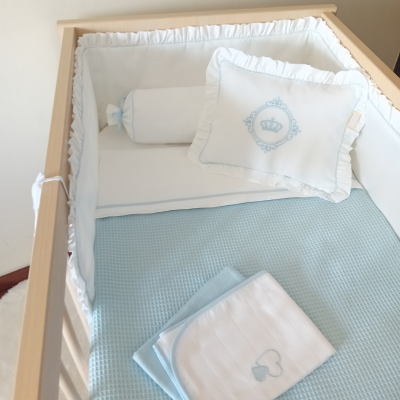 Berço de madeira com roupa de cama branca e azul claro com almofada bordada e mantas