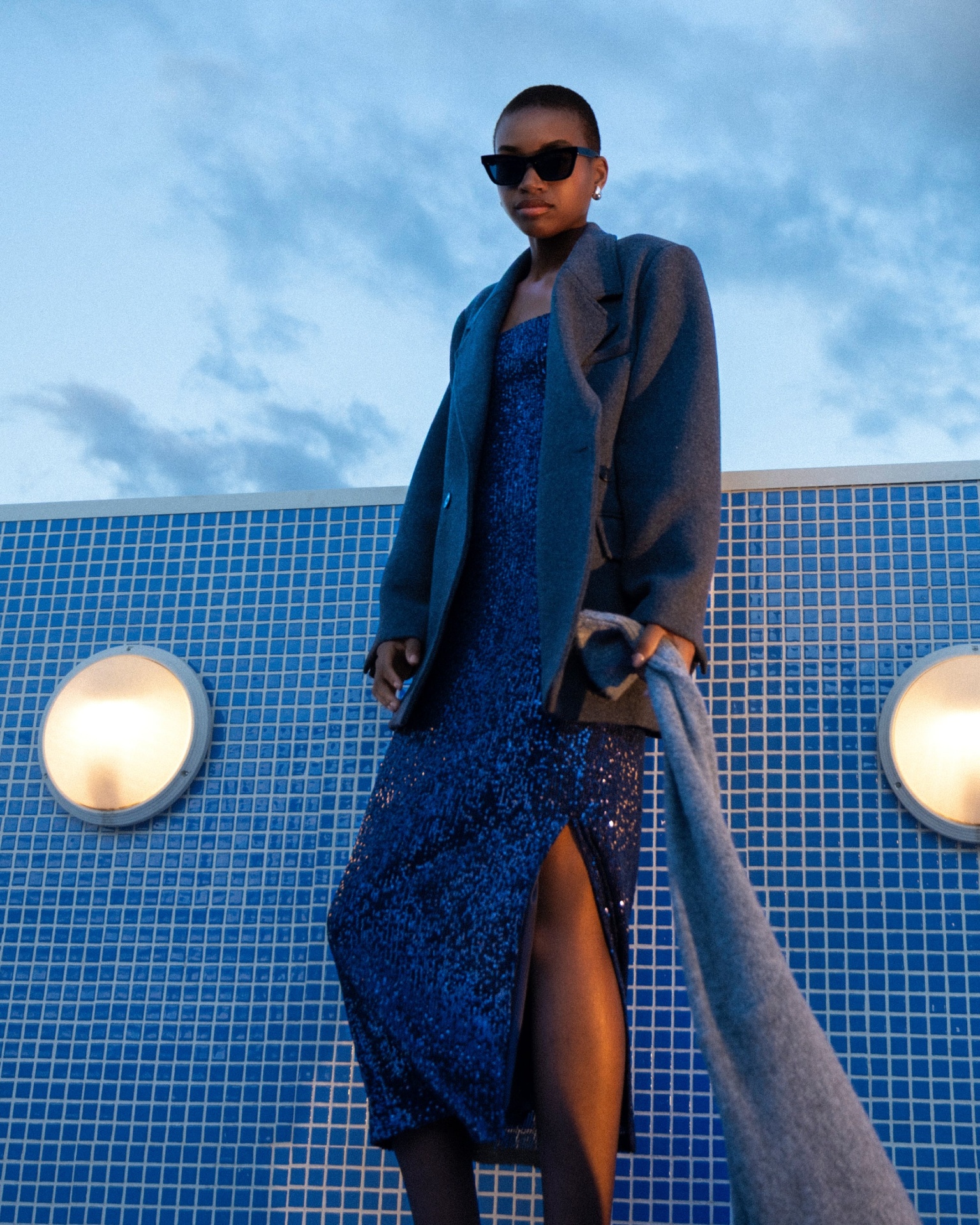 tori dress Mulher com vestido azul brilhante e casaco cinzento em frente a parede azul com azulejos