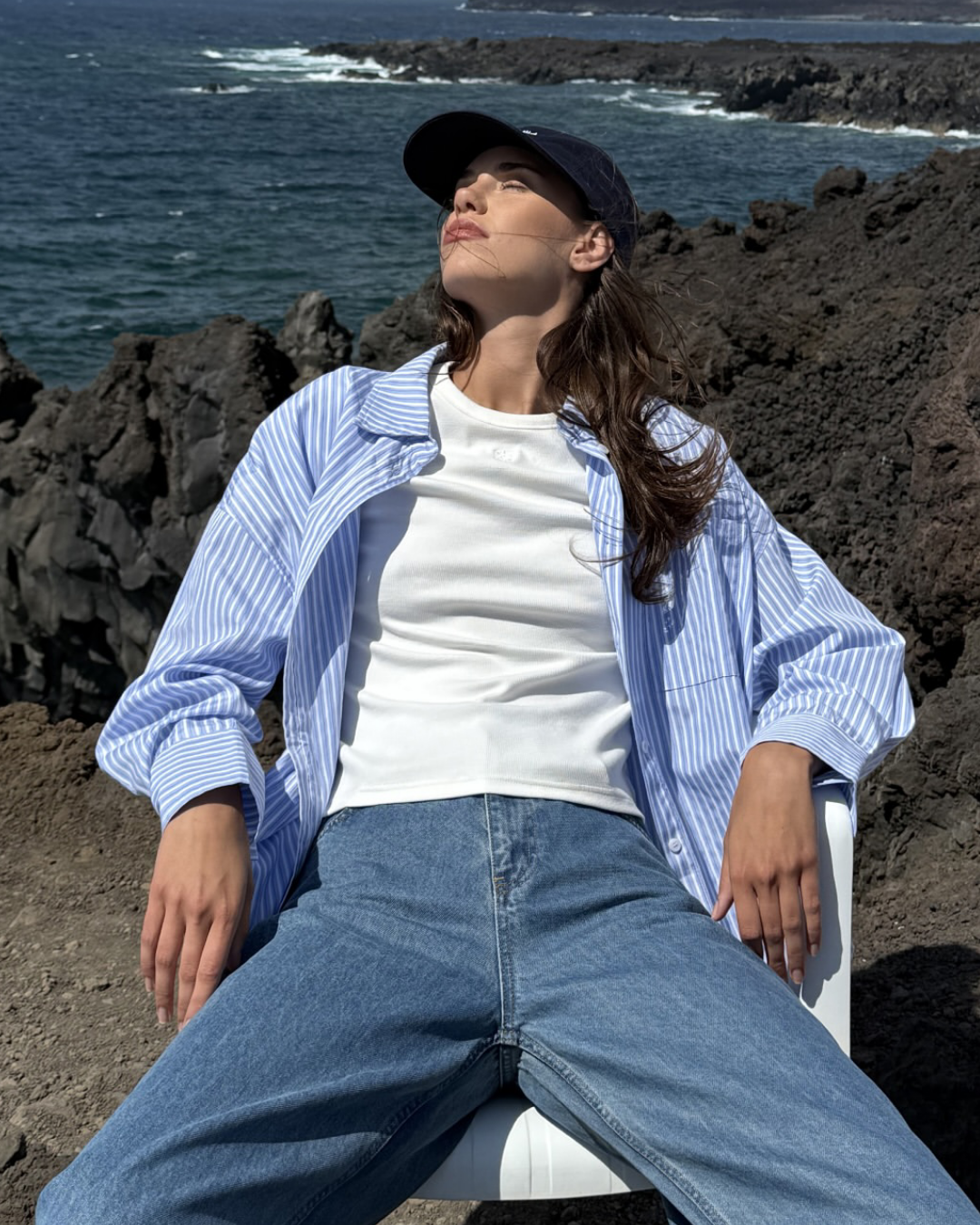 sure t-shirt Mulher sentada ao ar livre com camisa azul às riscas, t-shirt branca, jeans e boné preto.