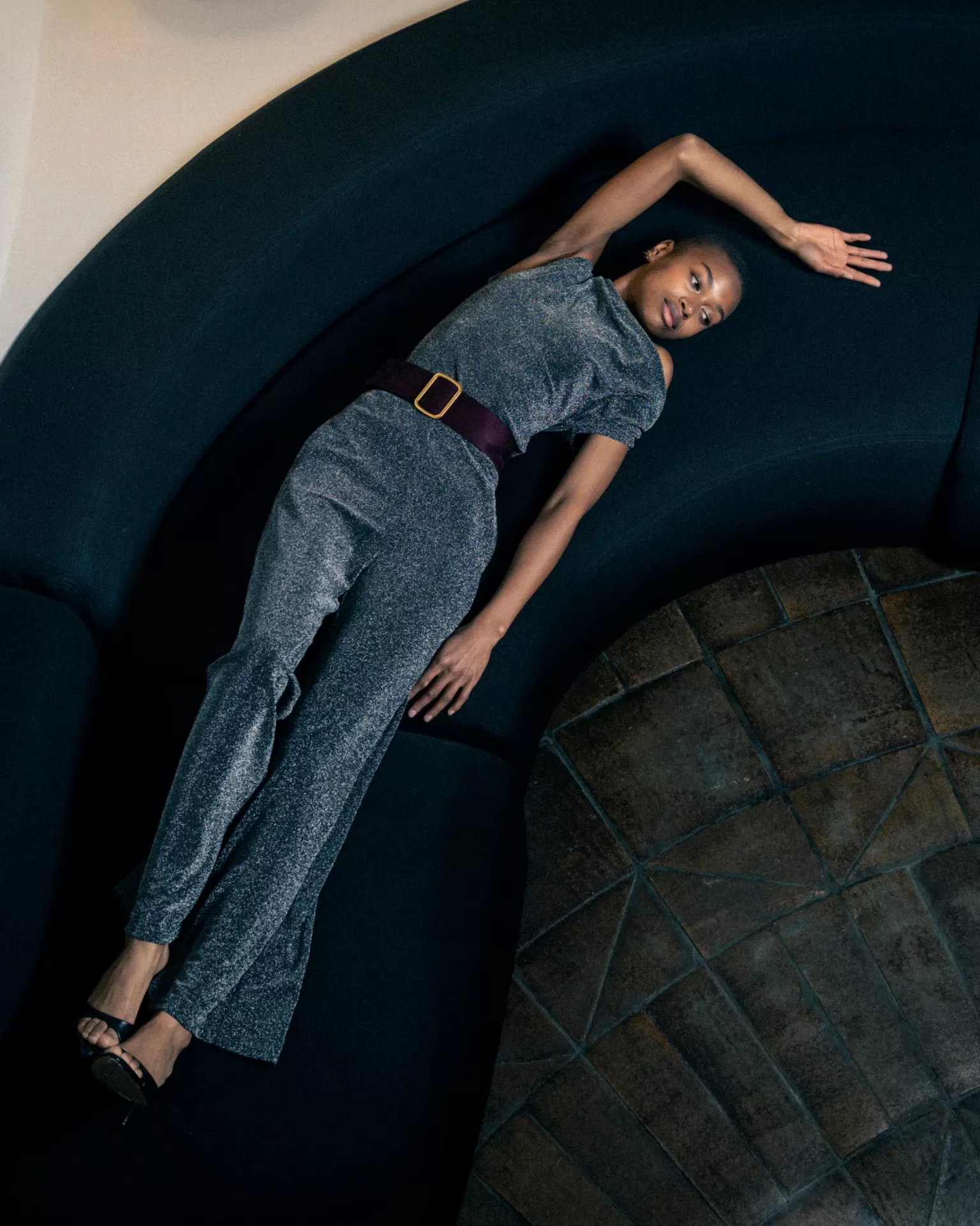 Macacão cinzento brilhante com cinto castanho em mulher num sofá preto curvo