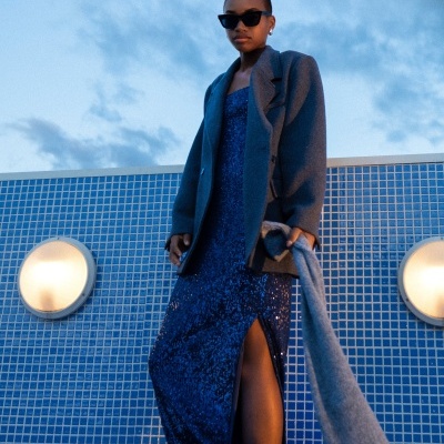 Mulher com vestido azul brilhante e casaco cinzento em frente a parede azul com azulejos