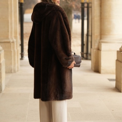 Mulher de costas com casaco de pêlo castanho escuro e calças brancas largas, segurando mala castanha pequena, em ambiente com colunas de pedra.