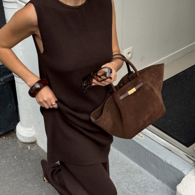 Mulher com vestido castanho escuro segurando mala castanha de camurça e óculos de sol.