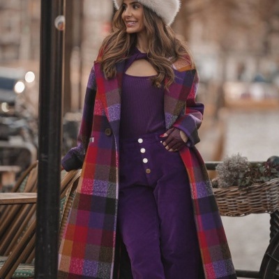 Mulher com casaco xadrez colorido, calças e camisola roxas, chapéu peludo creme em ambiente de cidade.