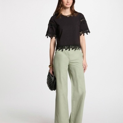 Mulher com top preto rendado, calças verdes largas, botas pretas e carteira preta em fundo branco