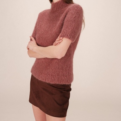 Mulher com camisola rosa felpuda de gola alta e saia castanha numa pose de braços cruzados