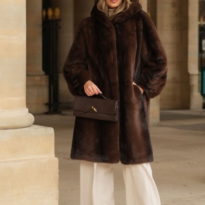 Mulher com casaco de pêlo castanho, calças brancas e mala castanha em área com colunas.