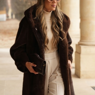 Mulher de casaco castanho escuro de pele sintética com capuz, óculos de sol rosa, calças creme e carteira preta em ambiente com pilares de pedra clara.