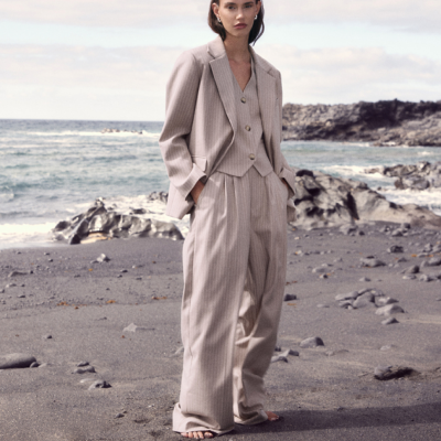 Fato bege claro com riscas finas composto por blazer, colete e calças largas, usado por modelo em praia rochosa.