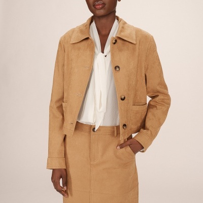 Conjunto feminino camel com casaco curto e saia, blusa branca e fundo neutro.